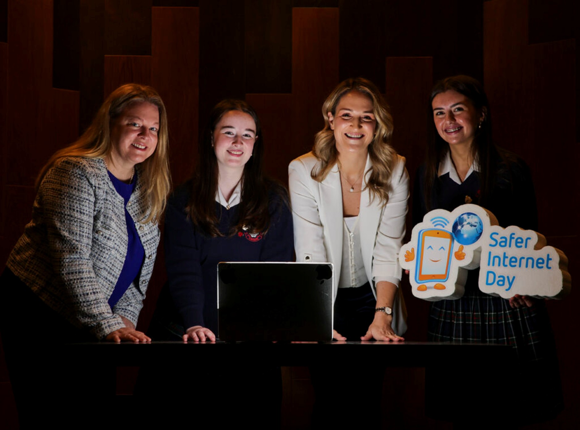 Four young women standing in front of an open laptop and holding a safer internet day banner
