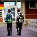Two young children facing away from the camera and walking towards their school with matching black and green backpacks.