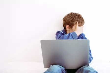 A scared young boy covering his eyes at the laptop screen