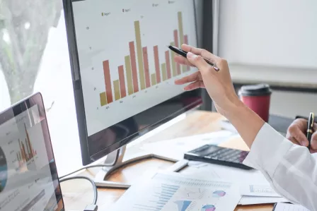 A hand holding a pen next to a PC screen showing a data sheet