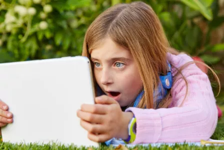 Child laying down in a park, looking at a tablet screen with a shocked expression
