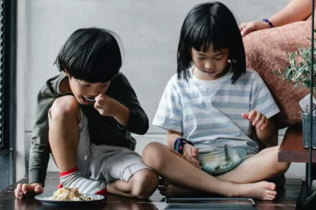 Two children sitting on the ground, eating and watching something on a tablet