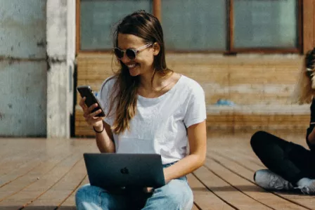 Young woman holding a laptop, smiling as she looks at her smartphone