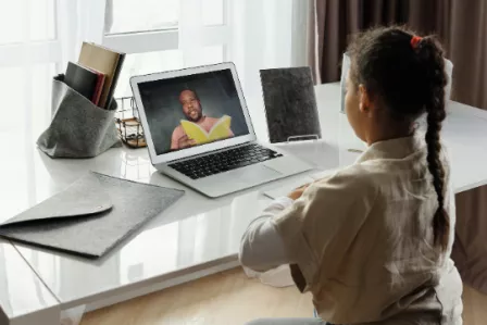 Girl sat at a desk using a laptop; a man on screen appears to be reading from a book