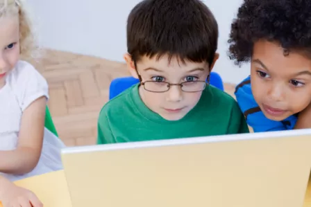 Three young children gathered around a laptop device, looking intently at the screen