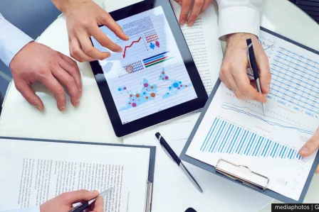 Three individuals (only hands visible) holding a tablet and a set of graphs on paper
