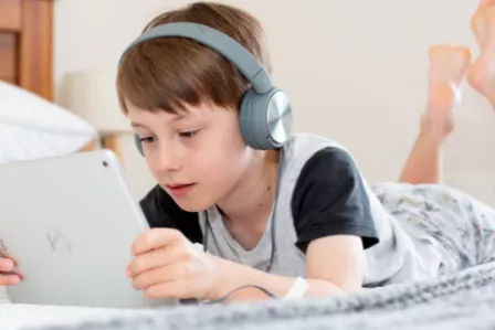 Child laying on a bed, with headphones on, watching content on a tablet
