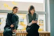 Two teenage girls are sitting on a bench by a large window. One of them is holding a smartphone and focusing on the screen, while the other leans over her shoulder, watching what the phone is showing.