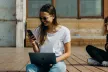 Young woman holding a laptop, smiling as she looks at her smartphone