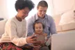 Two parents holding their toddler and showing them a laptop screen and a mobile phone.
