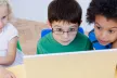 Three young children gathered around a laptop device, looking intently at the screen