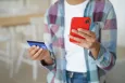A young person holding a red smartphone in one hand, and a credit card in the other hand