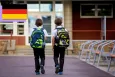 Two young children facing away from the camera and walking towards their school with matching black and green backpacks.